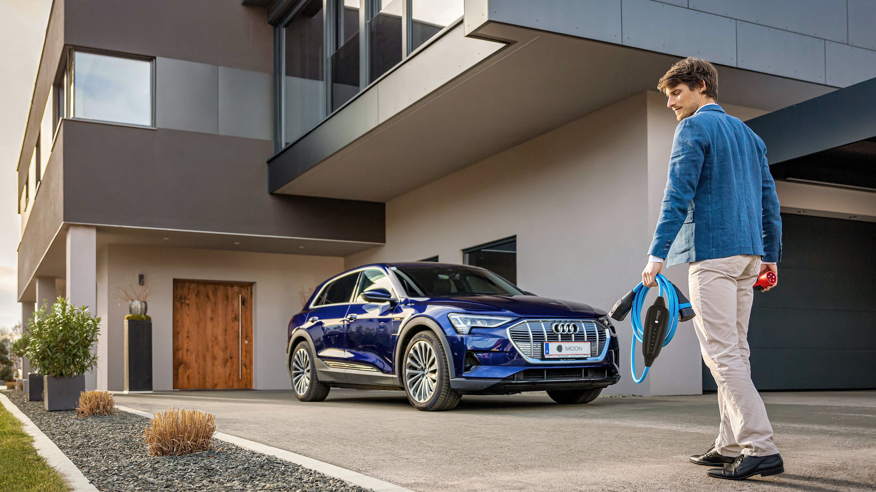 Man walks towards an electric car near a house with a cable in his hand